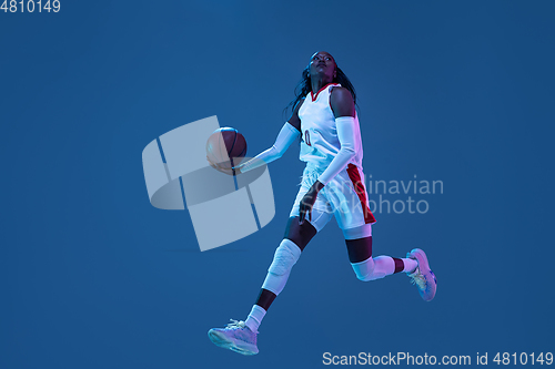 Image of Beautiful african-american female basketball player in motion and action in neon light on blue background. Concept of healthy lifestyle, professional sport, hobby.