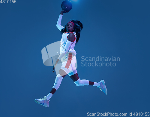 Image of Beautiful african-american female basketball player in motion and action in neon light on blue background. Concept of healthy lifestyle, professional sport, hobby.