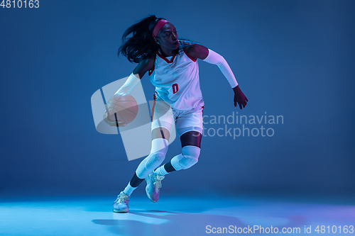 Image of Beautiful african-american female basketball player in motion and action in neon light on blue background. Concept of healthy lifestyle, professional sport, hobby.