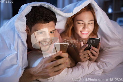 Image of happy couple using smartphones in bed at night