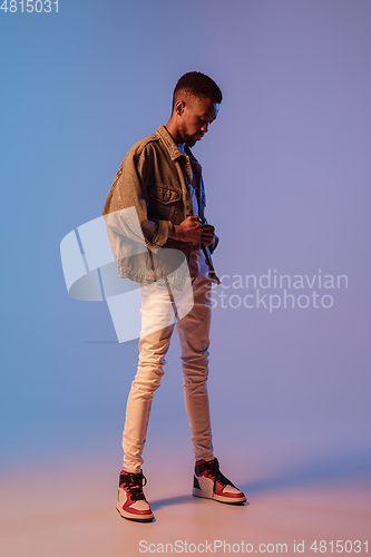 Image of Young stylish man in modern street style outfit isolated on gradient background in neon light. African-american fashionable model in look book, musician performing.