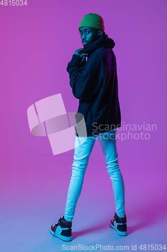 Image of Young stylish man in modern street style outfit isolated on gradient background in neon light. African-american fashionable model in look book, musician performing.