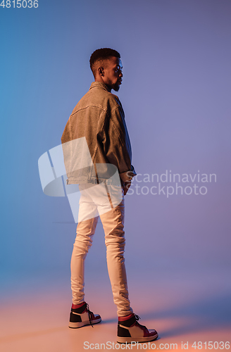 Image of Young stylish man in modern street style outfit isolated on gradient background in neon light. African-american fashionable model in look book, musician performing.