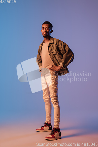 Image of Young stylish man in modern street style outfit isolated on gradient background in neon light. African-american fashionable model in look book, musician performing.