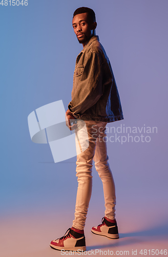 Image of Young stylish man in modern street style outfit isolated on gradient background in neon light. African-american fashionable model in look book, musician performing.