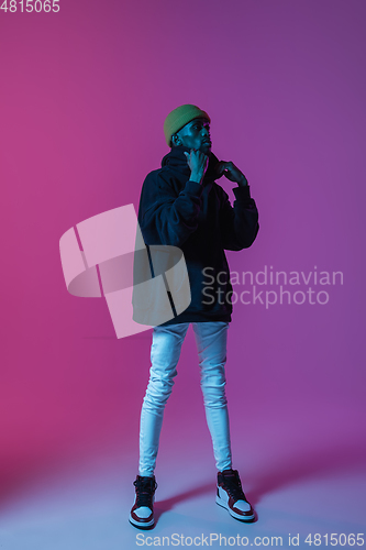 Image of Young stylish man in modern street style outfit isolated on gradient background in neon light. African-american fashionable model in look book, musician performing.