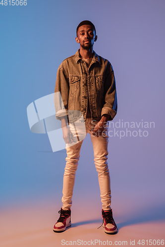 Image of Young stylish man in modern street style outfit isolated on gradient background in neon light. African-american fashionable model in look book, musician performing.