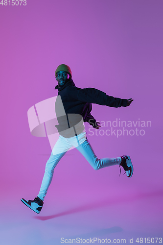 Image of Young stylish man in modern street style outfit isolated on gradient background in neon light. African-american fashionable model in look book, musician performing.