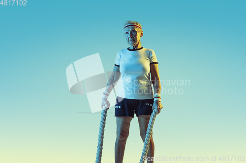 Image of Senior woman training with ropes in sportwear on gradient background in neon light