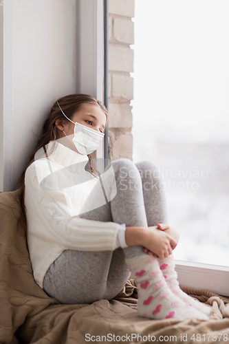 Image of sad girl in medical mask sitting on sill at home