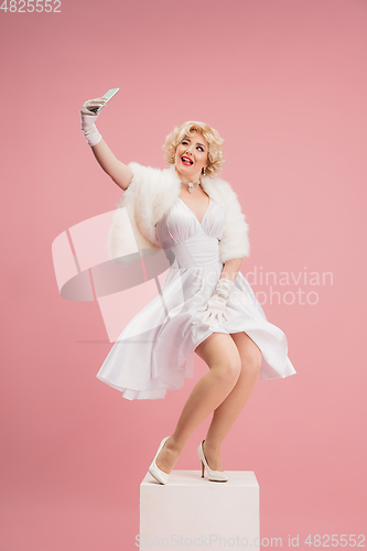 Image of Portrait of young woman in white dress on coral pink background. Female model as a legendary actress. Pin up. Concept of comparison of eras, modern, fashion, beauty.