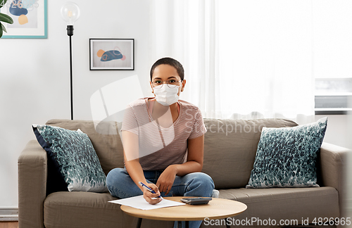 Image of woman in mask with papers and calculator at home