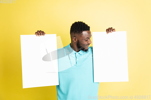 Image of Young African man with blank sheets of paper isolated over yellow studio background.