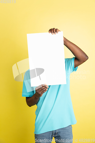 Image of Young African man with blank sheet of paper isolated over yellow studio background.