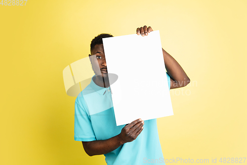 Image of Young African man with blank sheet of paper isolated over yellow studio background.