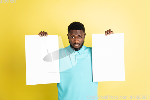 Image of Young African man with blank sheets of paper isolated over yellow studio background.