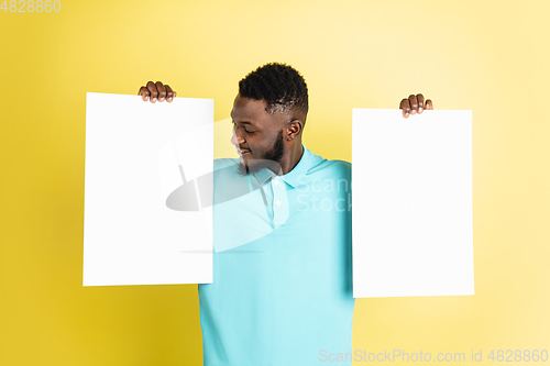 Image of Young African man with blank sheets of paper isolated over yellow studio background.