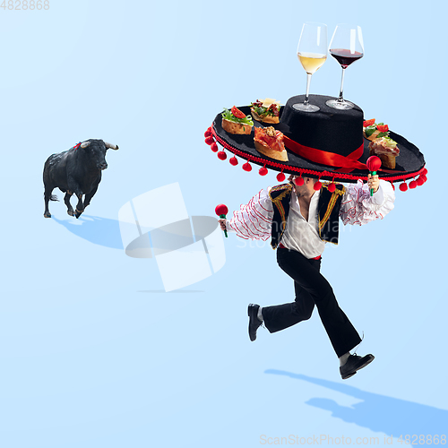 Image of Traditional corrida, bullfighter in sombrero with tapas and wine glasses is running away from bull on blue background