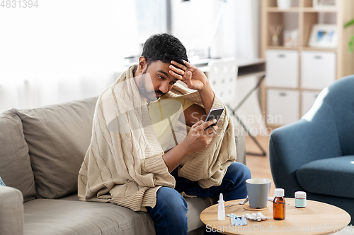 Image of sick young man in blanket with smartphone at home