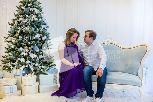 Image of Pregnant woman in an ultraviolet dress with husband sitting near Christmas tree