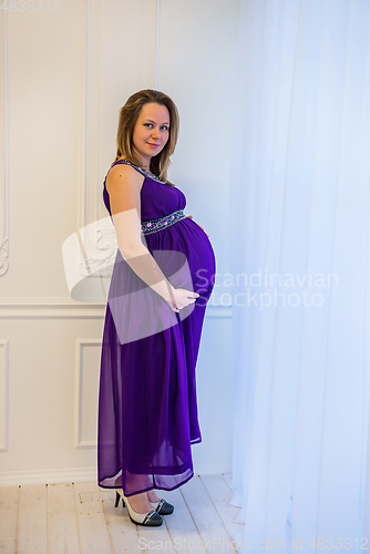 Image of Beautiful pregnant woman in an ultraviolet dress standing near the window