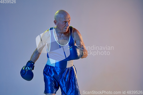 Image of Senior man wearing sportwear boxing isolated on gradient studio background in neon light. Concept of sport, activity, movement, wellbeing. Copyspace, ad.