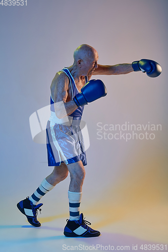 Image of Senior man wearing sportwear boxing isolated on gradient studio background in neon light. Concept of sport, activity, movement, wellbeing. Copyspace, ad.