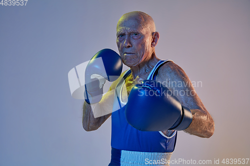 Image of Senior man wearing sportwear boxing isolated on gradient studio background in neon light. Concept of sport, activity, movement, wellbeing. Copyspace, ad.