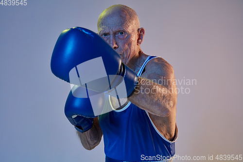 Image of Senior man wearing sportwear boxing isolated on gradient studio background in neon light. Concept of sport, activity, movement, wellbeing. Copyspace, ad.