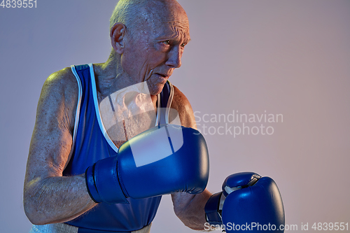 Image of Senior man wearing sportwear boxing isolated on gradient studio background in neon light. Concept of sport, activity, movement, wellbeing. Copyspace, ad.