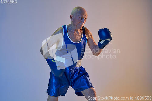 Image of Senior man wearing sportwear boxing isolated on gradient studio background in neon light. Concept of sport, activity, movement, wellbeing. Copyspace, ad.