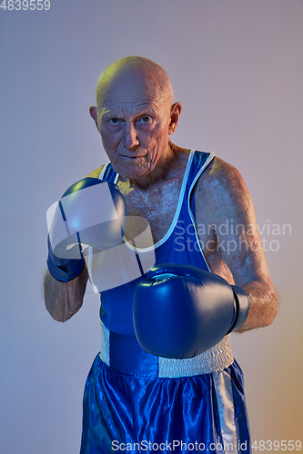 Image of Senior man wearing sportwear boxing isolated on gradient studio background in neon light. Concept of sport, activity, movement, wellbeing. Copyspace, ad.