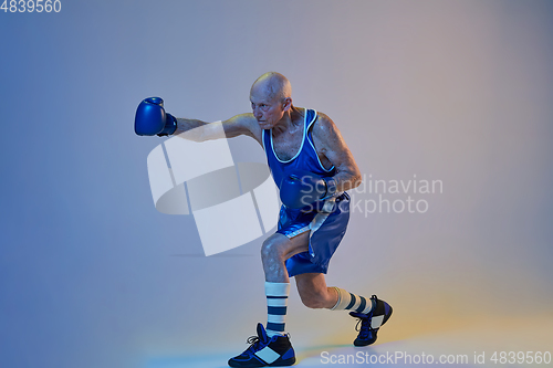 Image of Senior man wearing sportwear boxing isolated on gradient studio background in neon light. Concept of sport, activity, movement, wellbeing. Copyspace, ad.