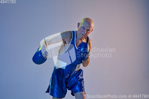 Image of Senior man wearing sportwear boxing isolated on gradient studio background in neon light. Concept of sport, activity, movement, wellbeing. Copyspace, ad.