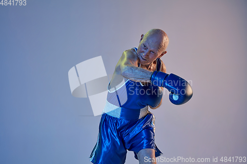 Image of Senior man wearing sportwear boxing isolated on gradient studio background in neon light. Concept of sport, activity, movement, wellbeing. Copyspace, ad.