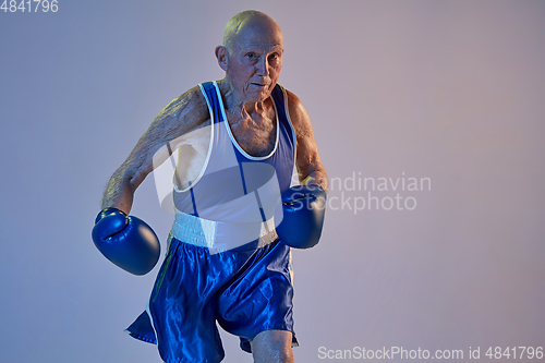 Image of Senior man wearing sportwear boxing isolated on gradient studio background in neon light. Concept of sport, activity, movement, wellbeing. Copyspace, ad.