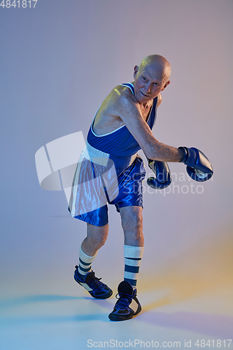 Image of Senior man wearing sportwear boxing isolated on gradient studio background in neon light. Concept of sport, activity, movement, wellbeing. Copyspace, ad.