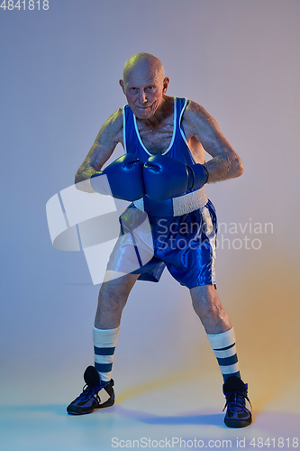 Image of Senior man wearing sportwear boxing isolated on gradient studio background in neon light. Concept of sport, activity, movement, wellbeing. Copyspace, ad.