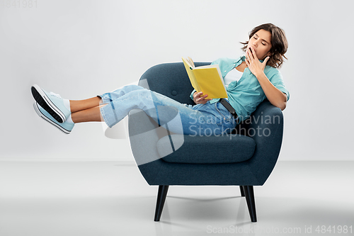 Image of bored woman in armchair reading book and yawning