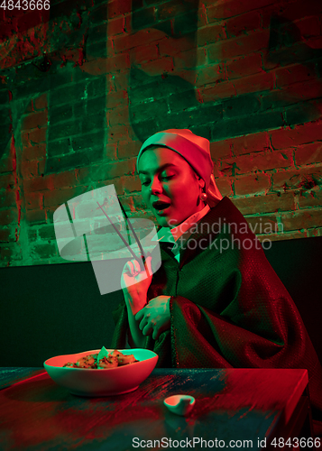 Image of Beautiful girl with a pearl earring taking lunch in modern cafe, restaurant in neon light