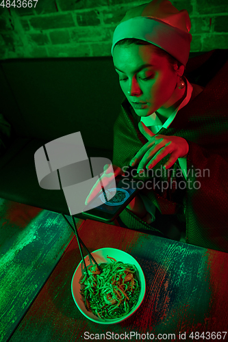 Image of Beautiful girl with a pearl earring taking lunch in modern cafe, restaurant in neon light