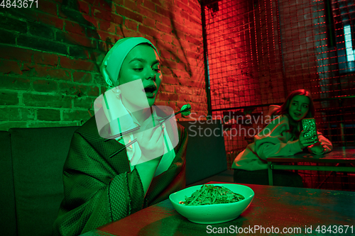 Image of Beautiful girl with a pearl earring taking lunch in modern cafe, restaurant in neon light