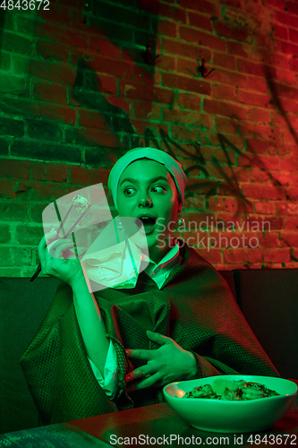 Image of Beautiful girl with a pearl earring taking lunch in modern cafe, restaurant in neon light