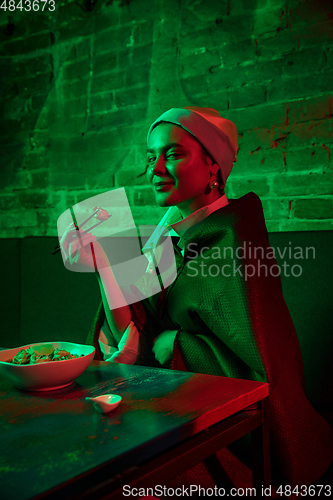 Image of Beautiful girl with a pearl earring taking lunch in modern cafe, restaurant in neon light