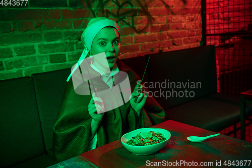 Image of Beautiful girl with a pearl earring taking lunch in modern cafe, restaurant in neon light