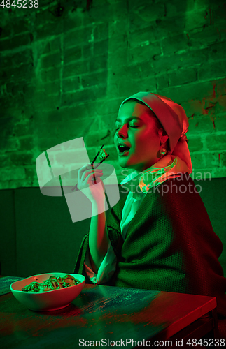 Image of Beautiful girl with a pearl earring taking lunch in modern cafe, restaurant in neon light