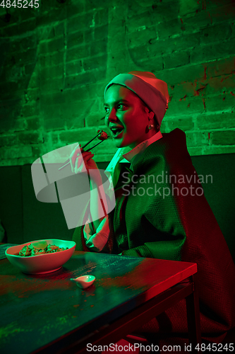 Image of Beautiful girl with a pearl earring taking lunch in modern cafe, restaurant in neon light