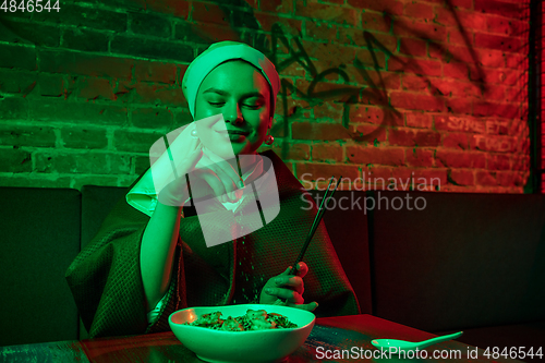 Image of Beautiful girl with a pearl earring taking lunch in modern cafe, restaurant in neon light