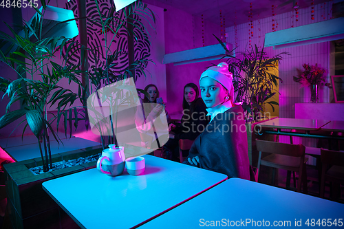 Image of Beautiful girl with a pearl earring taking lunch in modern cafe, restaurant in neon light