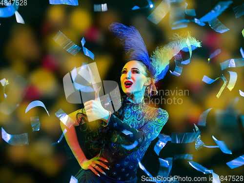 Image of Beautiful young woman in carnival and masquerade costume in colorful neon lights on black background in flying confetti